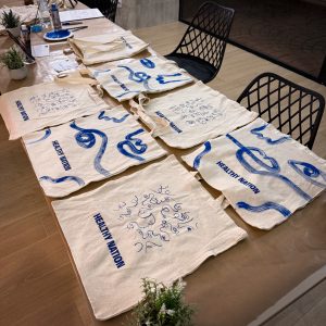 Tote bags de tela extendidas sobre una mesa de madera, personalizadas a mano con trazos artísticos y caligrafía en pintura azul con el texto Healthy Nation.