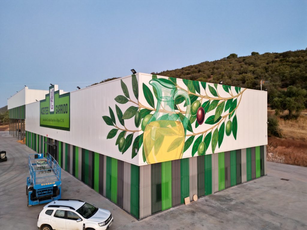 Mural corporativo pintado en una pared exterior para Aceites Garrido que muestra un diseño artístico de aceitunas y aceite, integrando la identidad visual de la marca en la fachada.